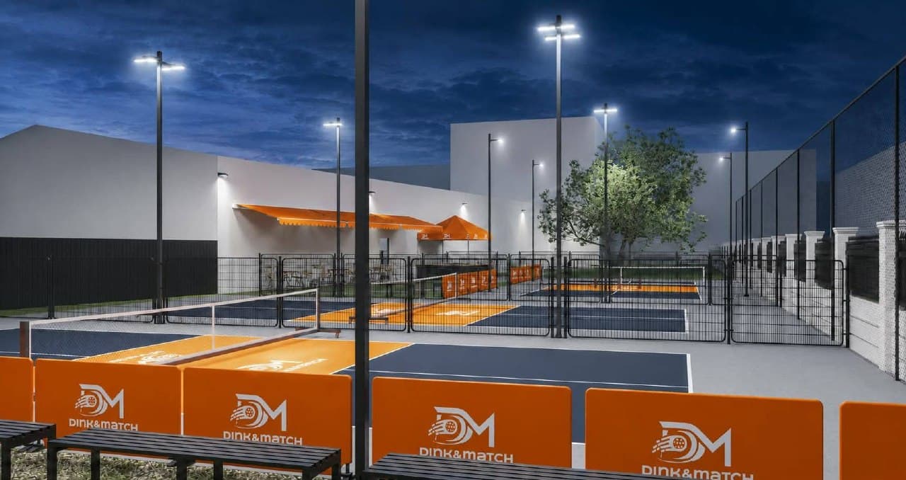 Outdoor pickleball courts at dusk under floodlights, with orange Dink&Match barriers along the near edge